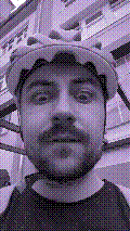 a selfie of a white person, wearing either headphones or a helmet, making an odd expression. the photo is edited to be colorized into a monochrome purple-ish color palette.